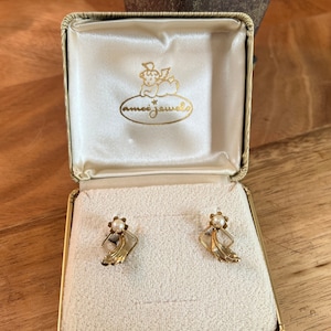 May include: A pair of gold earrings with a pearl accent, in a small, white box with the words "amoi jewels" printed on the lid.