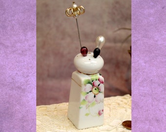 May include: A white porcelain pin cushion with a floral design in pink and green. The pin cushion has a decorative crown and pearl-topped pins. The base is rectangular, and the top is round with two pin holders.