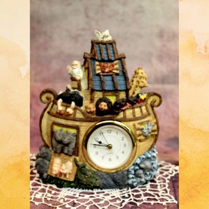 Può includere: Un orologio in ceramica a forma di arca di Noè con una colomba bianca in cima. L'arca è piena di animali, tra cui una giraffa, un leone e una zebra. Il quadrante dell'orologio è bianco con numeri e lancette neri.