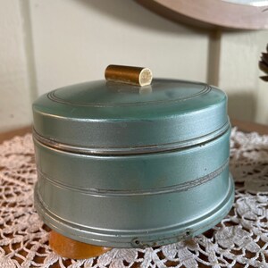 May include: A vintage, teal-colored, round music box with a brass knob on the lid. The music box is sitting on a white crocheted doily.