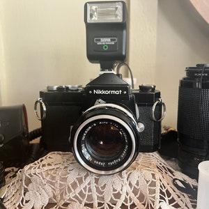May include: A black Nikon Nikkormat film camera with a silver lens and a flash attached. The camera is sitting on a white crocheted surface with two additional lenses.