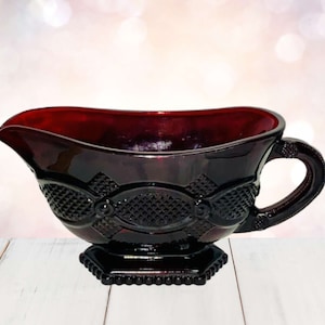 Könnte beinhalten: Eine rubinrote Glasschiffchen mit einem dekorativen Muster. Das Schüsselchen hat einen Ausguss, einen Griff und einen quadratischen Sockel. Das Glas hat eine glänzende Oberfläche und ist wahrscheinlich Vintage.