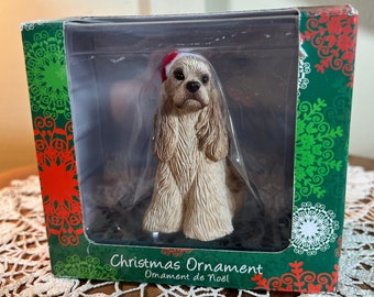 May include: A white and brown Cocker Spaniel dog wearing a red and white Santa hat, sitting on a black base. The dog is a Christmas ornament with the text "Christmas Ornament Ornament de Noel!" on the box.