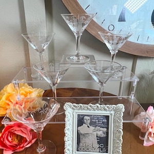 May include: Five clear glass martini glasses with etched designs are displayed on a two-tiered acrylic stand. The stand is on a wooden surface with flowers and a framed sign that reads "Mark of the New York Bar Family (Since 1921) ABC Mixing Cocktails".