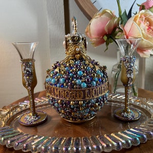 K&ouml;nnte beinhalten: Ein dekoratives Tablett mit zwei goldger&auml;nderten Gl&auml;sern und einer mit Juwelen besetzten Flasche. Die Flasche ist mit blauen, violetten und goldenen Perlen verziert und hat eine goldene Krone oben. Das Tablett hat eine schillernde Oberfl&auml;che.