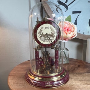 Puede incluir: Un reloj de péndulo vintage, dorado, con una esfera roja y una cúpula de vidrio. La esfera del reloj presenta una escena de una pareja en un jardín con el texto "Lieb's" encima. El reloj está sentado en una mesa de madera.