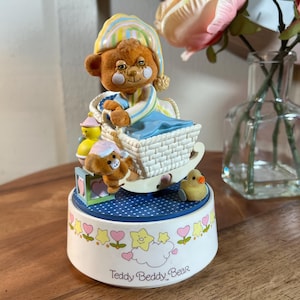 Puede incluir: Un carrusel musical blanco con un oso de peluche que lleva una gorra de dormir azul y amarilla. El oso está en una cuna de mimbre con una manta azul. Hay dos osos de peluche más pequeños y un pájaro amarillo en el carrusel. La base del carrusel está decorada con corazones rosas, estrellas amarillas y el texto "Teddy Beddy Bear".