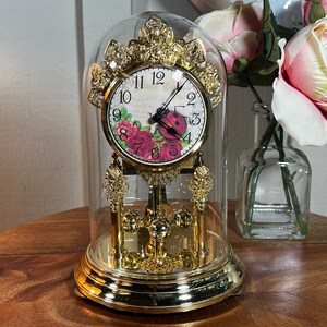 May include: A gold-colored decorative clock with a glass dome. The clock face features a floral design with a ladybug. The clock is on a wooden surface, with a vase of flowers in the background.