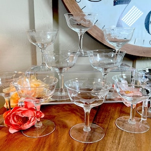 Op de afbeelding: Een set van negen heldere glazen champagnecoupes met gegraveerde ontwerpen. De coupes zijn gestapeld op een heldere acryl-trapstandaard. De coupes zijn in een verspringend patroon gerangschikt, met drie coupes op de bovenste rij, drie coupes op de middelste rij en drie coupes op de onderste rij.
