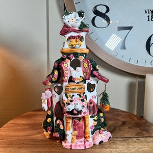 Puede incluir: Un portavelas de cerámica con forma de iglesia con un exterior rosa y blanco. La iglesia tiene un letrero que dice "Old Stone Church" y está decorada con flores y corazones.