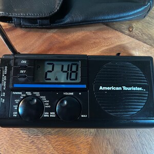 May include: Black American Tourister digital clock radio with a telescoping antenna. The display shows 2:48. Features include AM/FM radio, alarm, and volume control. A black leather case is in the background.