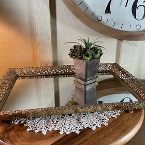 Könnte beinhalten: Ein verziertes Tablett in Goldtönen mit einer verspiegelten Oberfläche und einem kunstvollen, geprägten Design. Eine kleine Sukkulente in einem Topf steht auf dem Tablett.