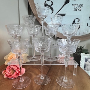 May include: A collection of clear, etched glass champagne coupe glasses. Each glass features a delicate floral design on the bowl and base, with a slender stem. The glasses are arranged on a clear acrylic stand, with a vintage clock in the background.