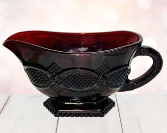 Op de afbeelding: Een robijnrode glazen juskom met een decoratief patroon. De juskom heeft een tuit, een handvat en een vierkante basis. Het glas heeft een glanzende afwerking en is waarschijnlijk vintage.