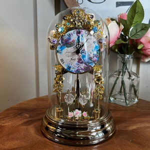 May include: A decorative clock with a floral design under a glass dome. The clock face features a blue and white floral pattern with Roman numerals. The clock is gold-colored with crystal accents and a pendulum. A vase of pink roses is in the background.