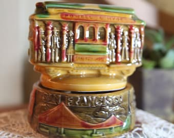 May include: A ceramic music box shaped like a San Francisco cable car with the words "Powell & Hyde" and "San Francisco" on it. The cable car is painted in red, yellow, green, and brown with figures of people on the sides.