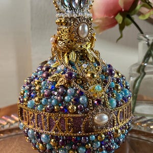 K&ouml;nnte beinhalten: Eine runde, violette und blaue Glasflasche mit einer goldenen Krone oben. Die Flasche ist mit Perlen und Juwelen bedeckt, und das Wort "DELUXE" ist in goldenen Buchstaben um die Mitte geschrieben.