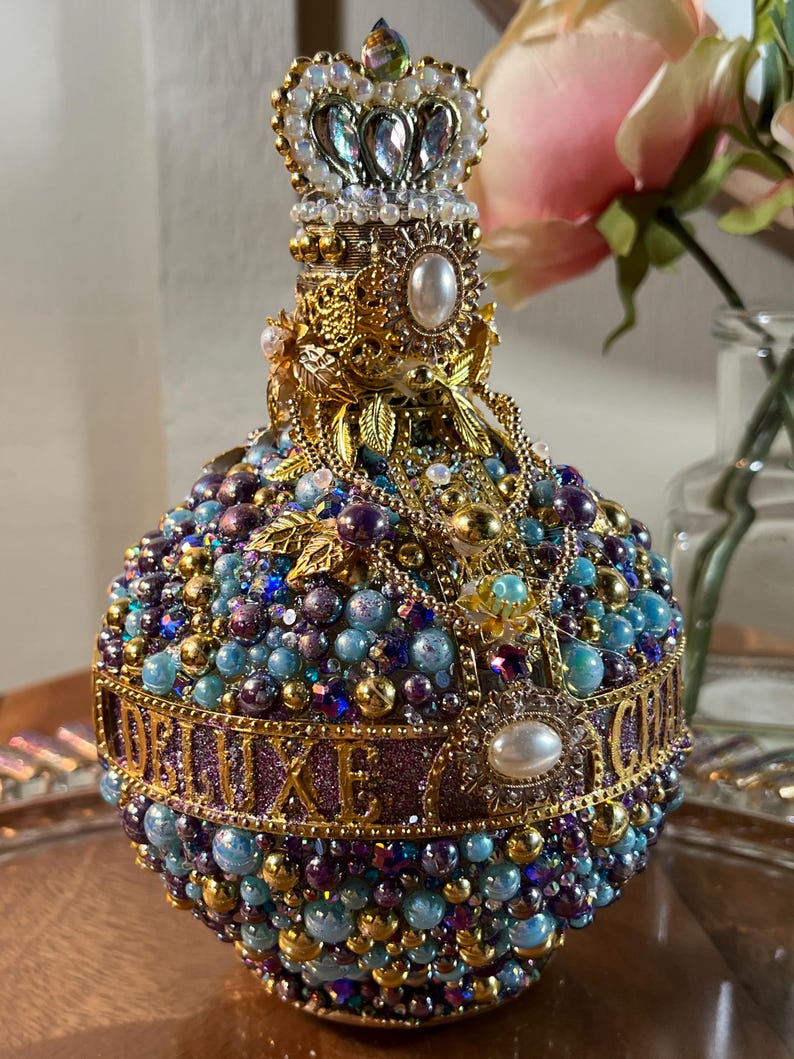 K&ouml;nnte beinhalten: Eine runde, violette und blaue Glasflasche mit einer goldenen Krone oben. Die Flasche ist mit Perlen und Juwelen bedeckt, und das Wort "DELUXE" ist in goldenen Buchstaben um die Mitte geschrieben.