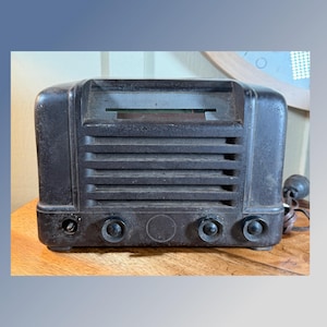Puede incluir: Una radio vintage de color marrón oscuro con forma rectangular. La radio tiene listones horizontales en la parte delantera y varios botones en la parte inferior. Un dial es visible detrás de una ventana rectangular. Un cable y un pequeño botón redondo están en el lado derecho.