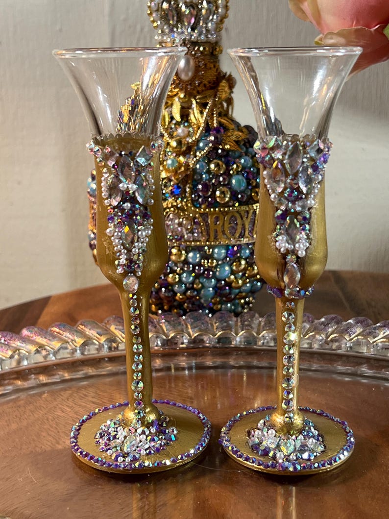 K&ouml;nnte beinhalten: Zwei goldbemalte Schnapsgl&auml;ser mit violetten und klaren Strasssteinen. Die Gl&auml;ser stehen auf einem goldenen Tablett.