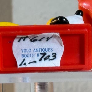 May include: A close-up of a red rectangular object with a white label that reads "VOLO ANTIQUES BOOTH # 703". The object is partially obscuring a yellow and black object. The background is a light color.
