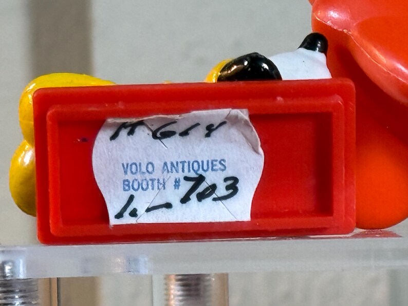 May include: A close-up of a red rectangular object with a white label that reads "VOLO ANTIQUES BOOTH # 703". The object is partially obscuring a yellow and black object. The background is a light color.