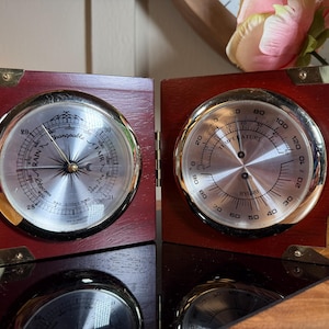 Puede incluir: Una estación meteorológica vintage con un barómetro y un termómetro. Los instrumentos están enmarcados en un marco de madera oscura con esquinas de latón. El dial del barómetro indica "Fair" y "Rain". El termómetro muestra la temperatura y la humedad.