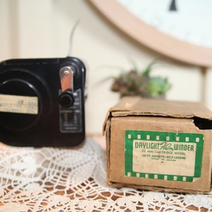 Puede incluir: Un enrollador de película negro con una manivela y una etiqueta que dice "35 MM CARTRIDGE MODEL 100 FT. CAPACITY - SELF-LOCKING" está sentado en un encaje blanco de ganchillo junto a una caja de cartón marrón con una etiqueta verde que dice "DAYLIGHT Film Winder 35 MM CARTRIDGE MODEL 100 FT. CAPACITY - SELF-LOCKING".