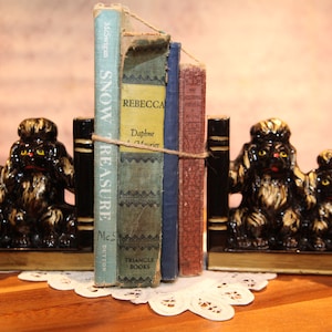 May include: A set of black and white poodle bookends holding a stack of vintage books. The books are titled "Snow Treasure", "Rebecca", "Daphne & Maurice", and "Triangle Books".