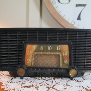 May include: A vintage black and gold Sylvania radio with a dial that reads from 55 to 16. The radio has a black grille and two gold knobs.