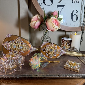 May include: An ornate vanity set with a vintage aesthetic. The set includes two hand mirrors, a small jewelry box, and a decorative rose. The mirrors and box are embellished with pearls, rhinestones, and gold accents. A candle and flowers complete the display.