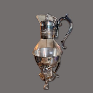 Könnte beinhalten: Eine Weinkaraffe aus klarem Glas und versilbertem Metall mit bauchigem Glaskörper, einem silbernen Kragen mit Gittermuster, einem geschwungenen verzierten Griff und einem dreibeinigen Fuß mit drei detaillierten Füßen unter der silbernen Schale.