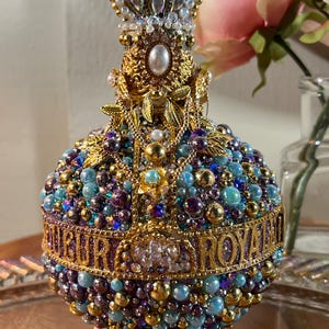 K&ouml;nnte beinhalten: Eine goldene und violette, mit Juwelen besetzte Flasche mit einer Krone oben. Die Flasche ist mit Perlen und Juwelen verziert und tr&auml;gt die Aufschrift "Royal".