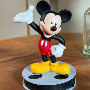 May include: A Mickey Mouse figurine, featuring black ears, a black body, red shorts, and yellow shoes. The figure is mounted on a grey film reel base. Mickey's white gloved hand is raised. A classic Disney collectible.