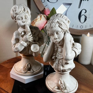 Puede incluir: Dos figuritas de cerámica blanca, una de un niño tocando el saxofón y la otra de una niña con las manos sobre las orejas. Ambas figuritas están sobre bases blancas con detalles florales.