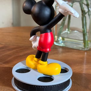 May include: A Mickey Mouse figurine in a classic pose, wearing red shorts, yellow shoes, and white gloves. The black figure stands on a light blue film reel base, a nostalgic nod to cinema.