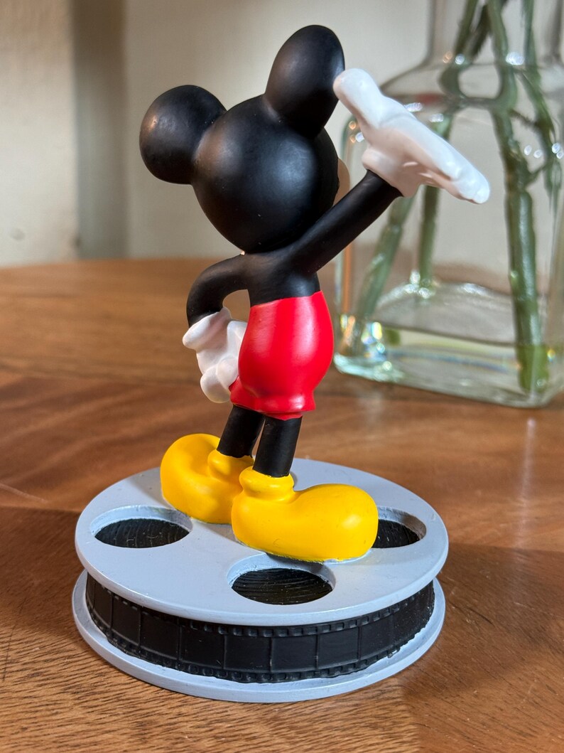 May include: A Mickey Mouse figurine in a classic pose, wearing red shorts, yellow shoes, and white gloves. The black figure stands on a light blue film reel base, a nostalgic nod to cinema.