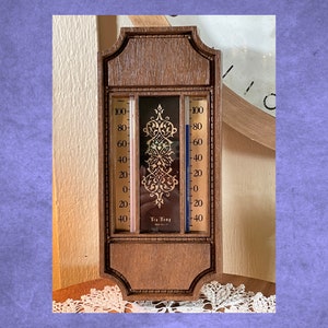 Puede incluir: Un termómetro de interior/exterior con marco de madera y diseño dorado. El termómetro tiene dos escalas, una para Fahrenheit y otra para Celsius. El termómetro indica "Tru Temp" en el centro.