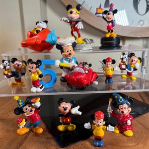 May include: A collection of Mickey Mouse figurines in various poses and outfits. The figurines are on clear acrylic shelves. Some figurines are holding birthday cakes, and others are in different costumes. A red airplane and the number 5 are also present.