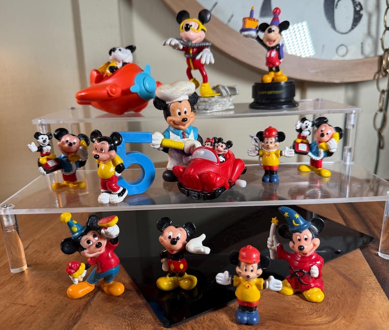 May include: A collection of Mickey Mouse figurines in various poses and outfits. The figurines are on clear acrylic shelves. Some figurines are holding birthday cakes, and others are in different costumes. A red airplane and the number 5 are also present.