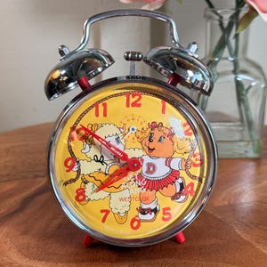 May include: A vintage Westclox alarm clock with a yellow face featuring cartoon characters. The clock has a chrome finish, red accents, and a dual bell design. The clock's face displays the time with red hands and numerals.