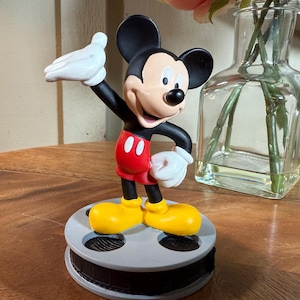 May include: A Mickey Mouse figurine in a classic pose, with black ears, red shorts, yellow shoes, and white gloves. The figure is mounted on a film reel base. A clear glass vase with pink flowers is in the background.