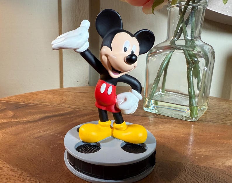 May include: A Mickey Mouse figurine in a classic pose, with black ears, red shorts, yellow shoes, and white gloves. The figure is mounted on a film reel base. A clear glass vase with pink flowers is in the background.