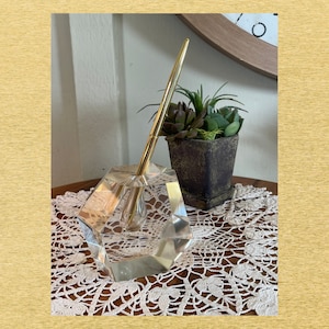 Puede incluir: Un tintero de cristal transparente y facetado con una pluma dorada insertada. El tintero tiene un diseño geométrico y está sobre una mesa de madera con un tapete de encaje blanco. Una pequeña planta suculenta en maceta está en el fondo.