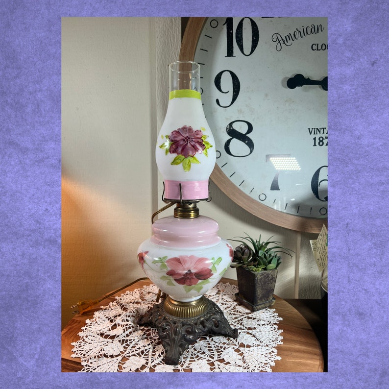 May include: A vintage white oil lamp with a pink base and a white shade with pink and green floral designs. The lamp has a metal base with ornate details and sits on a white lace doily.