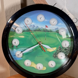 May include: A round wall clock with a black frame and a golf course scene on the face. White golf balls serve as hour markers, each labeled with a black number from 1 to 12. The clock features white hour and minute hands and a red second hand.