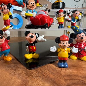 May include: A collection of Mickey Mouse figurines in various poses and outfits. The figurines are brightly colored, featuring red, yellow, blue, and black. Some are holding objects, and one is in a red car. The figurines are displayed on a shelf.