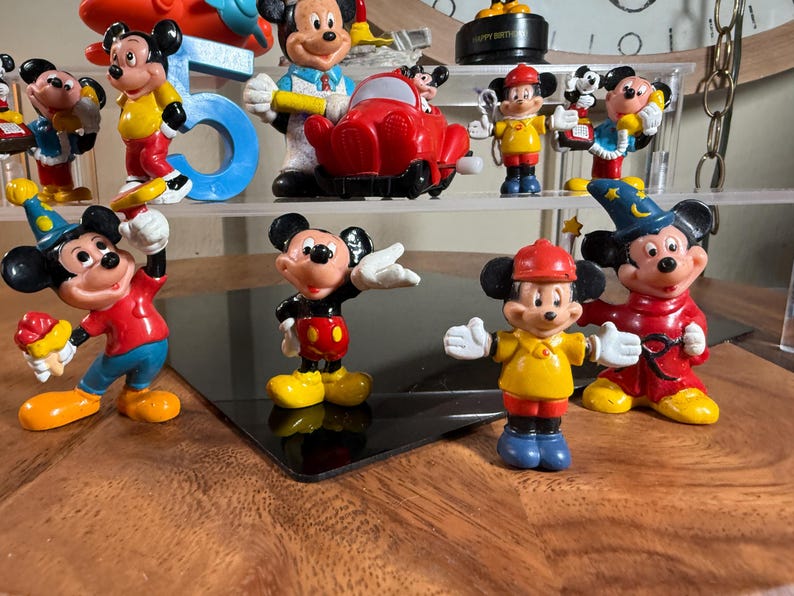 May include: A collection of Mickey Mouse figurines in various poses and outfits. The figurines are brightly colored, featuring red, yellow, blue, and black. Some are holding objects, and one is in a red car. The figurines are displayed on a shelf.