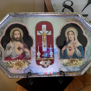 May include: An octagonal religious display featuring images of Jesus, a crucifix, and Mary. The images are framed with ornate gold and pink accents, set against a silver background. The display is supported by a black metal stand.