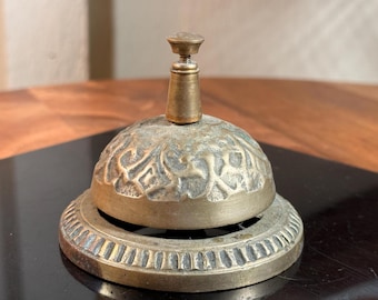 May include: Antique brass bell with a decorative floral pattern on the dome. The bell has a round base and a small handle on top. The metal has a weathered appearance, suggesting age and use. A classic item for home decor or a vintage collection.